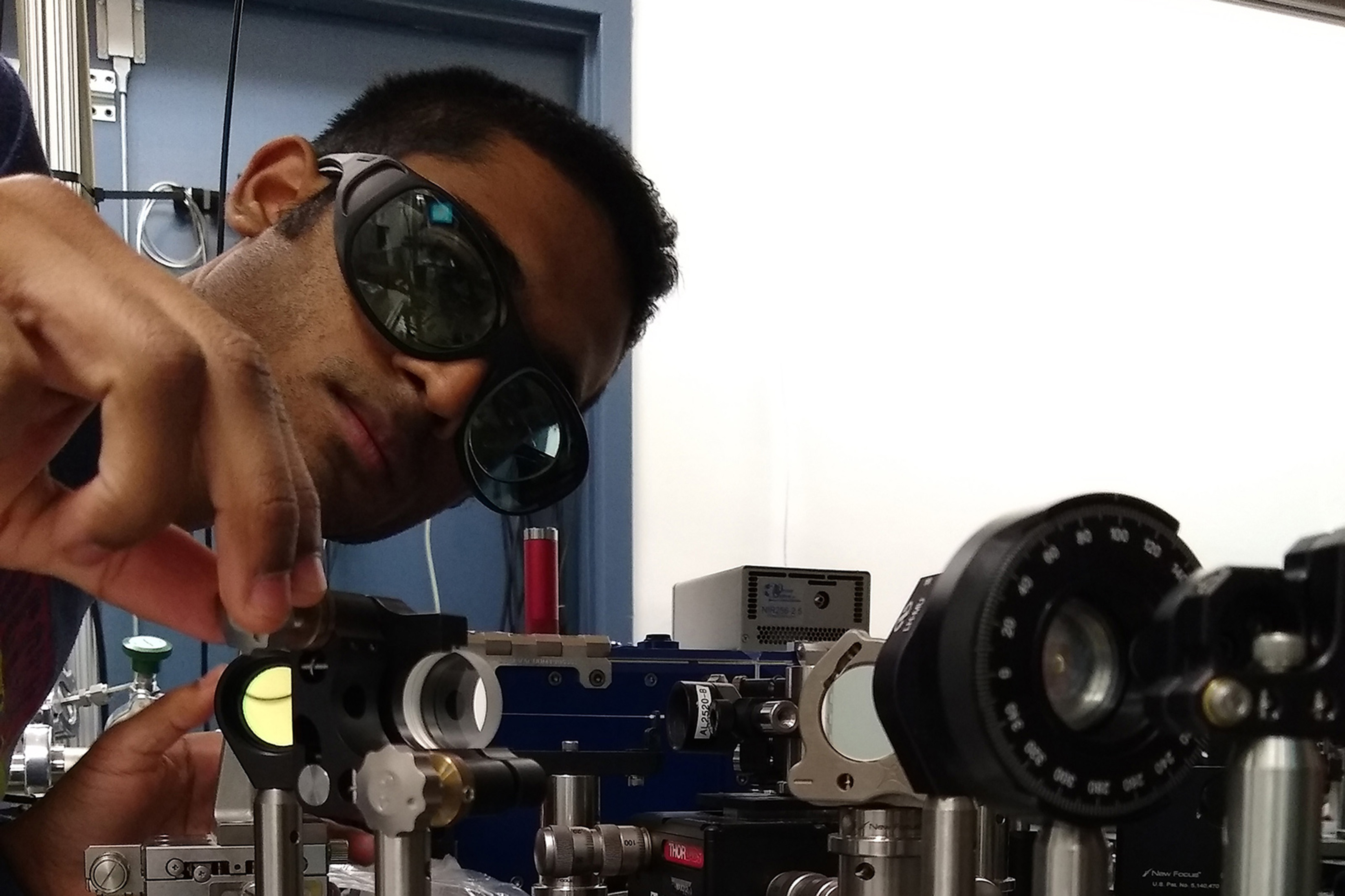 Sahil Pontula is shown in dark glasses leaning over some equipment that includes an array of lenses and various metal parts. He appears to be adjusting some element of the machinery.