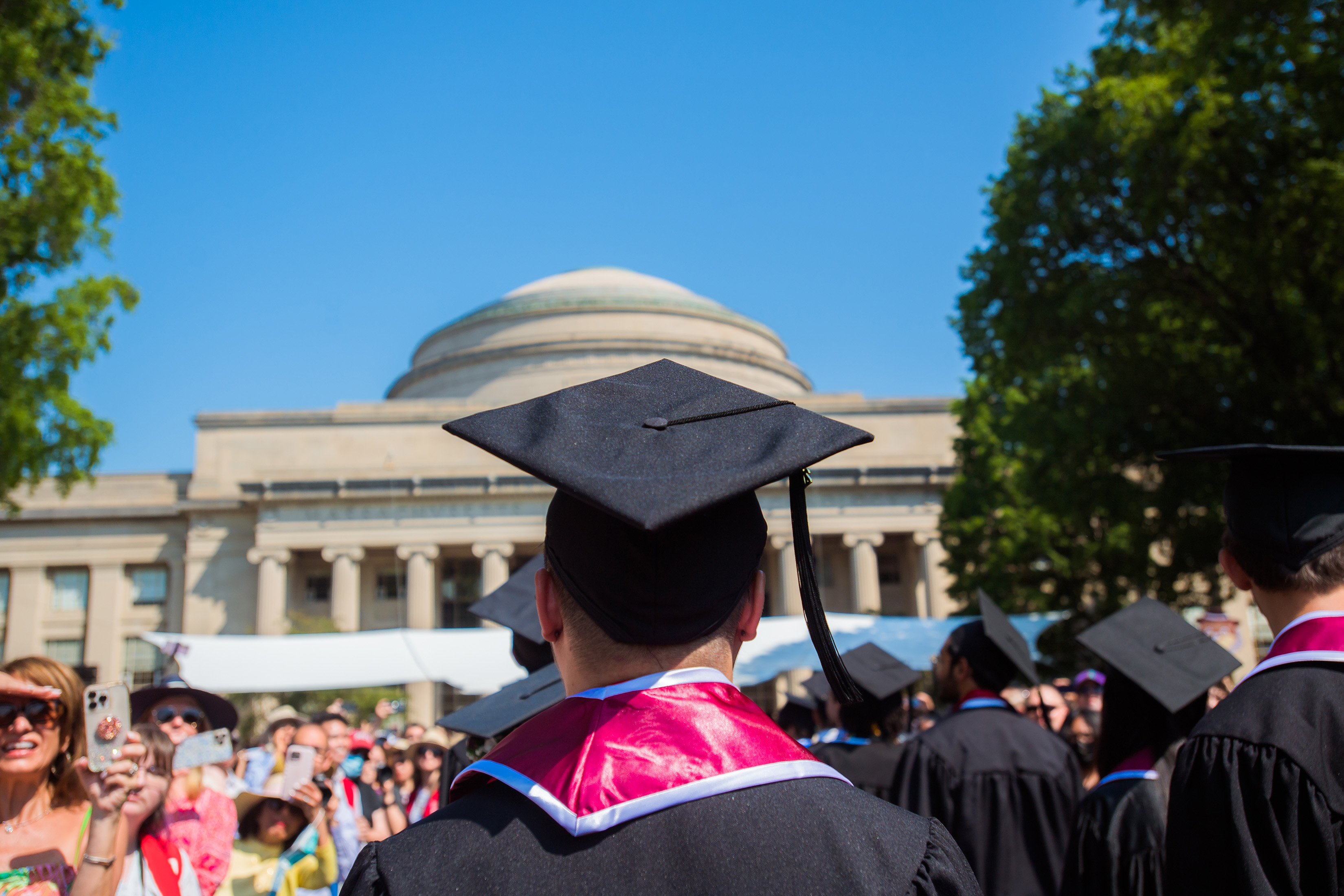 A person in a black graduation cap is shown from the back, from the shoulders up. In the background is the MIT dome, and there are more graduates on the right and people with cameras on the left.