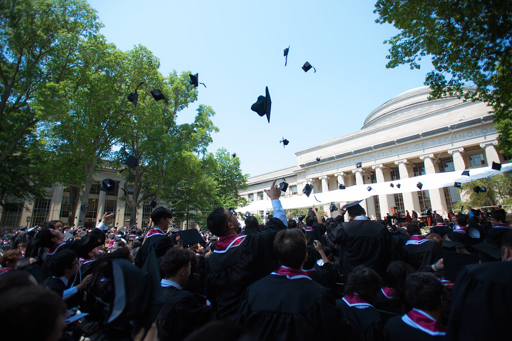 A photo of MIT's commencement, with rows of students sitting with their backs facing the camera in graduation caps and gowns outside with MIT's Building 10 in the background while students throw their caps in the air
