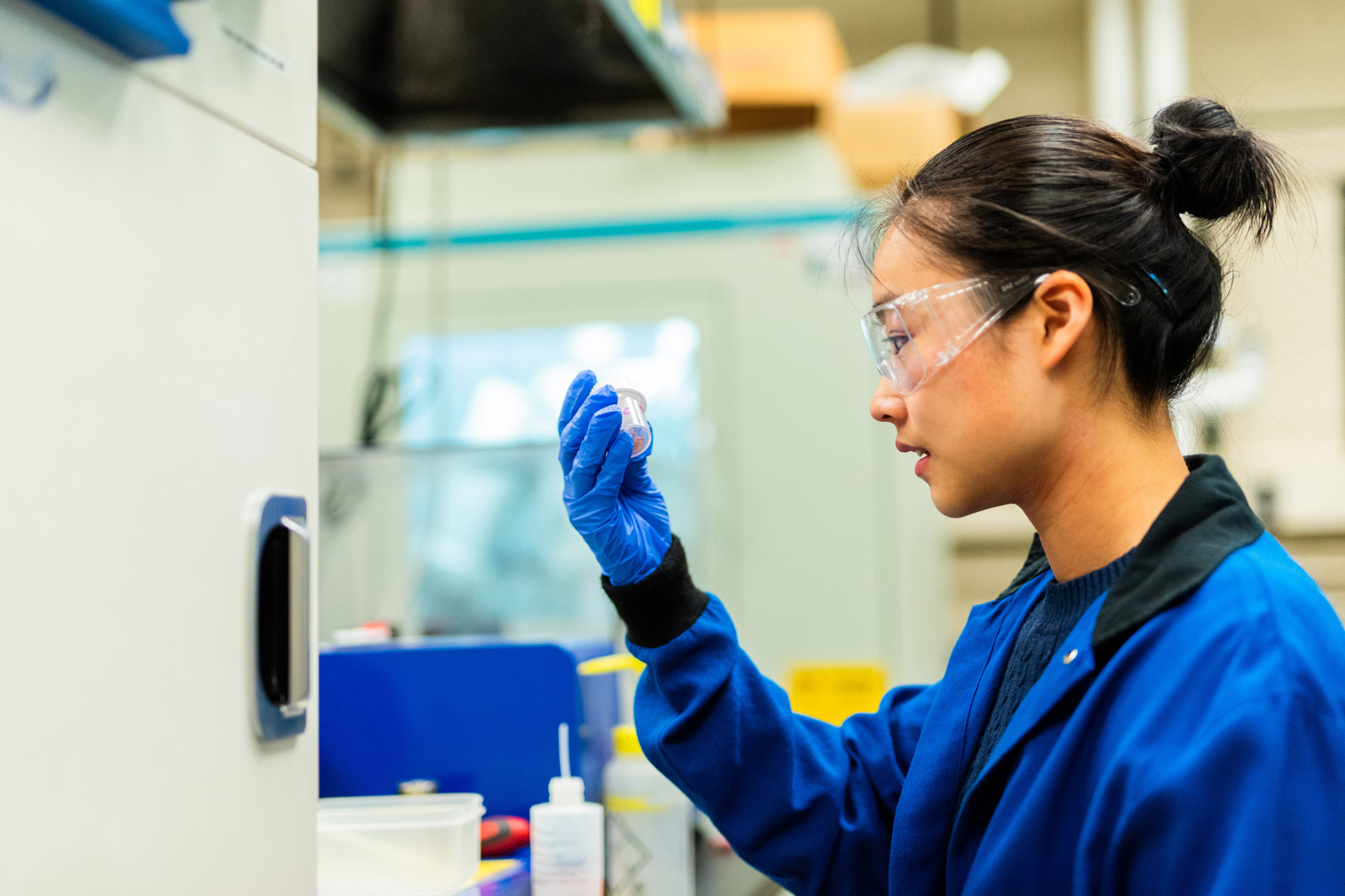 Shu Yang Zhang is shown in a lab setting. She is wearing goggles and looking at a small jar that she holds in one gloved hand. 