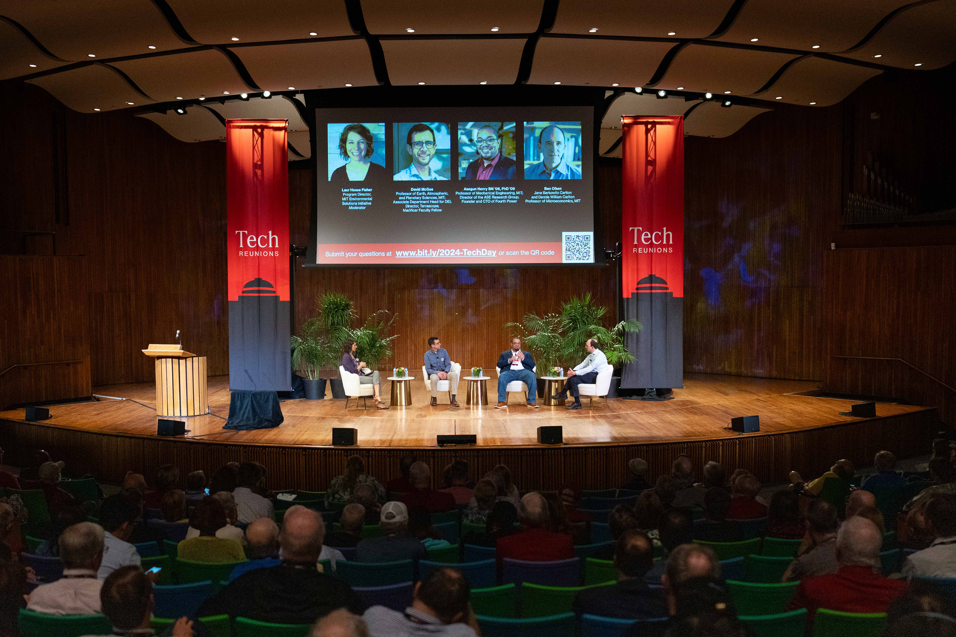 A view of a stage from the audience shows four people on stage. A large projection shows four headshots and the screen is framed by large red banners with the words Tech Reunions on them.
