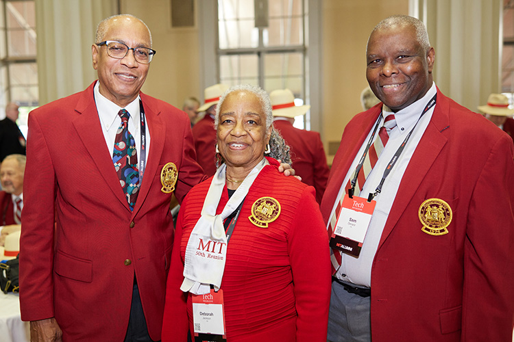 Two men and one woman pose together wearing red jackets and other MIT paraphernalia. The woman is in the center.