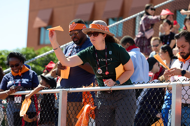 A woman in a straw hat decorated with an orange bandana stands poised to throw an orange paper airplane from behind a short chain-link fence. Others in the stands behind her are also wearing orange bandanas and holding orange paper airplanes.