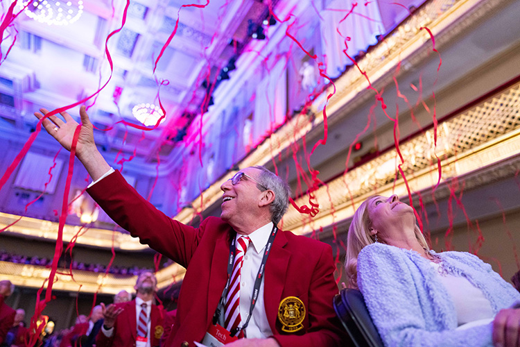 A man in a red jacket and MIT tie holds his hand out to catch red streamers cascading down inside a theater. A woman next to him looks up. Two tiers of balconies can be seen behind them.