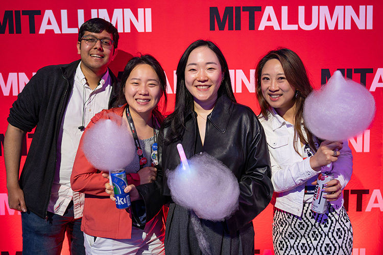 Four people pose in front of a background that says MIT ALUMNI. Three hold cotton candy.