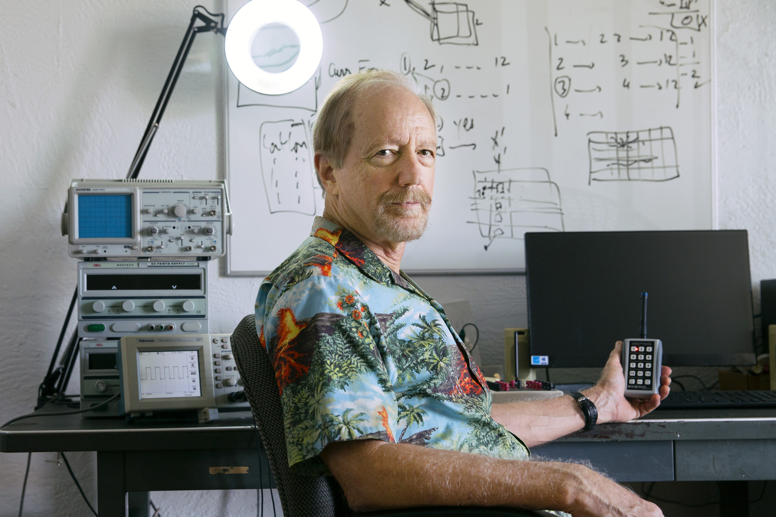 Charlie Holdaway is shown from elbows up, sitting at a desk and holding a keypad that has an antenna. Behind him on the left, machinery and a circular lamp can be seen. On the right, there is a computer screen; Behind him, there is a whiteboard with some numbers and figures visible.