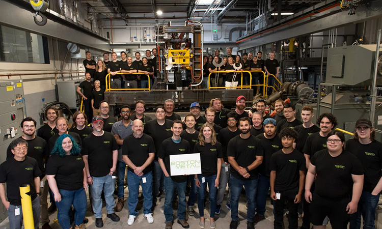 A huge group of people poses in an industrial setting. Everyone is wearing a black T-shirt with a green logo. In the center, one person holds a sign reading "Boston Metal."