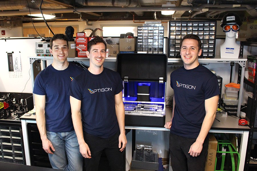 Three men in navy T-shirts (two shirts clearly read Optigon) pose in a workshop. On the workbench between two of them is an open black case. Parts inside appear blue and gray.