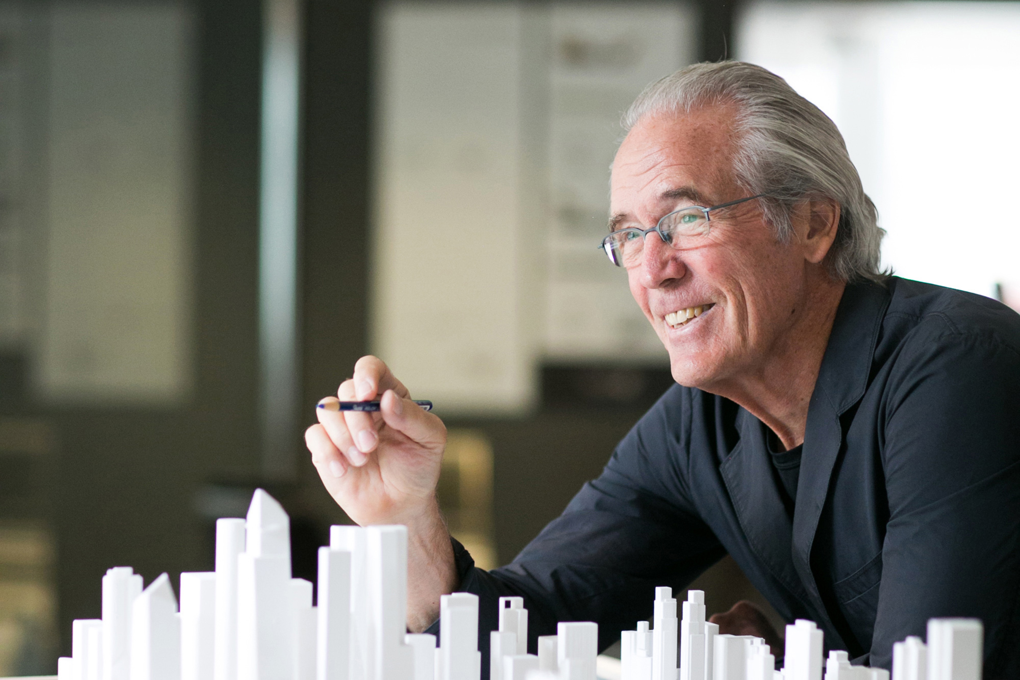 William Pedersen is shown from chest up holding a pen in one hand and leaning over an array of white model buildings.