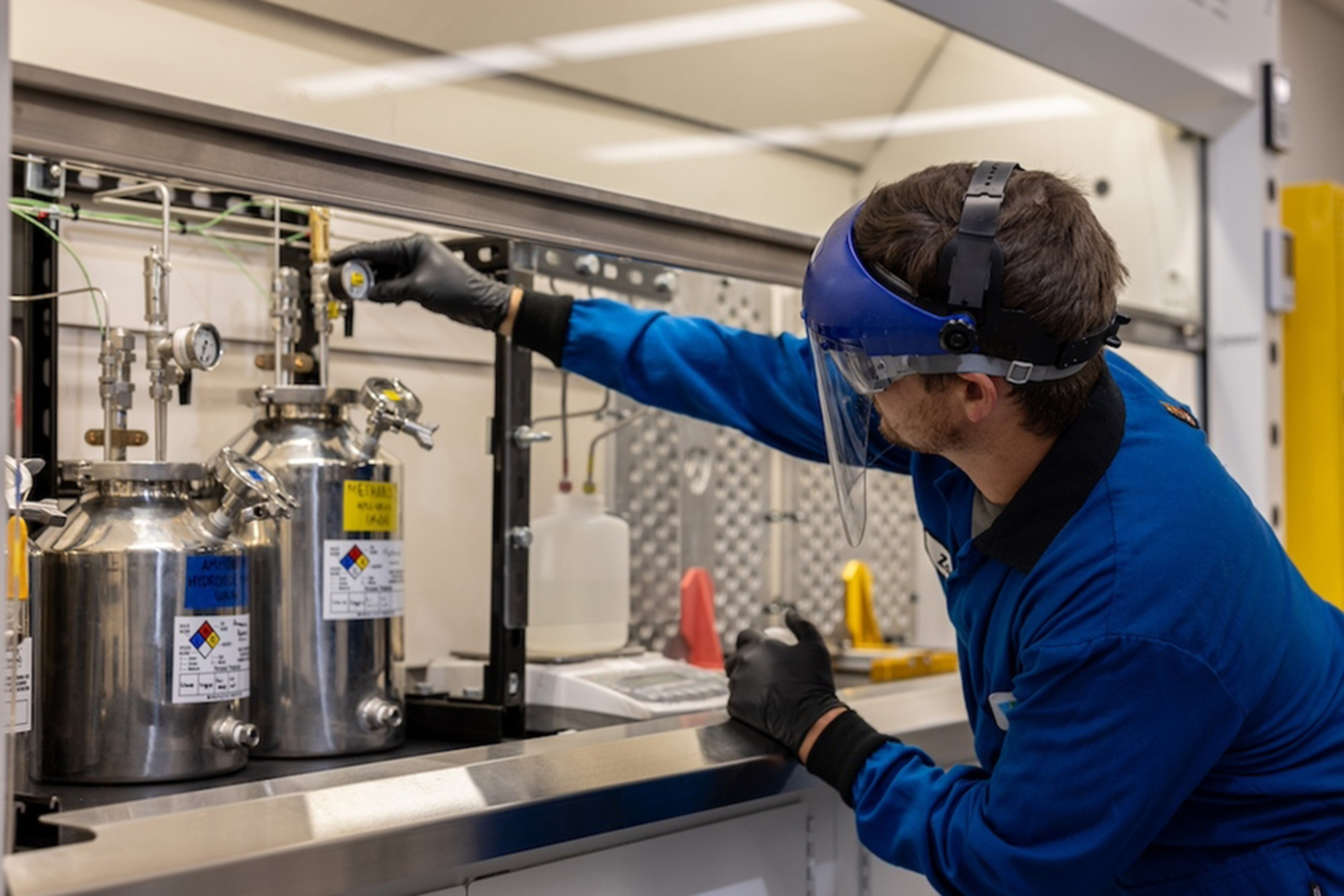 Zach Goodman, wearing full lab protection, adjusts a gauge above a stainless steel cylinder in a lab.