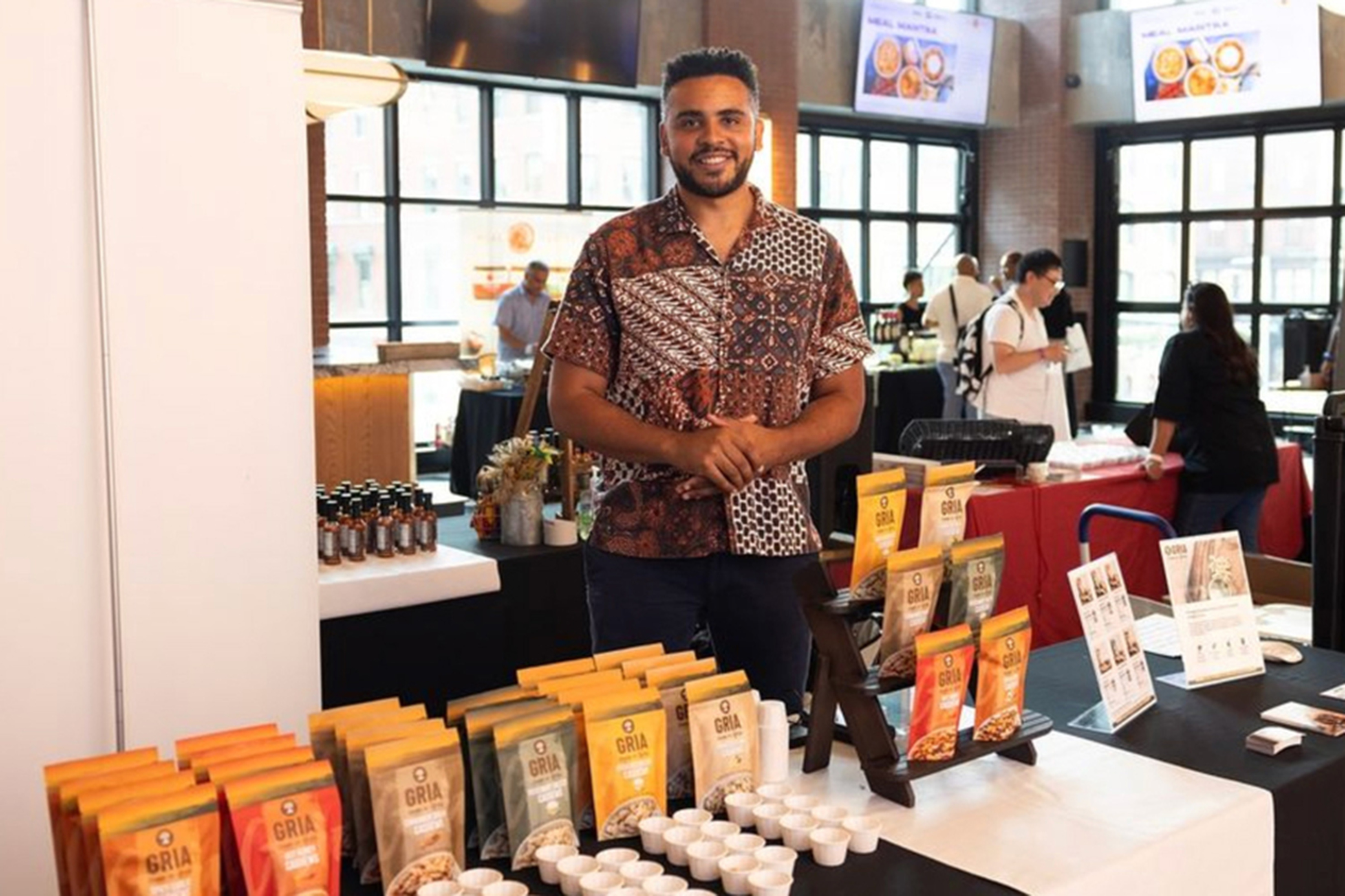 Joshua Reed-Diawuoh stands behind a table featuring GRIA products and small cups. Other tables and shoppers are visible in the background.