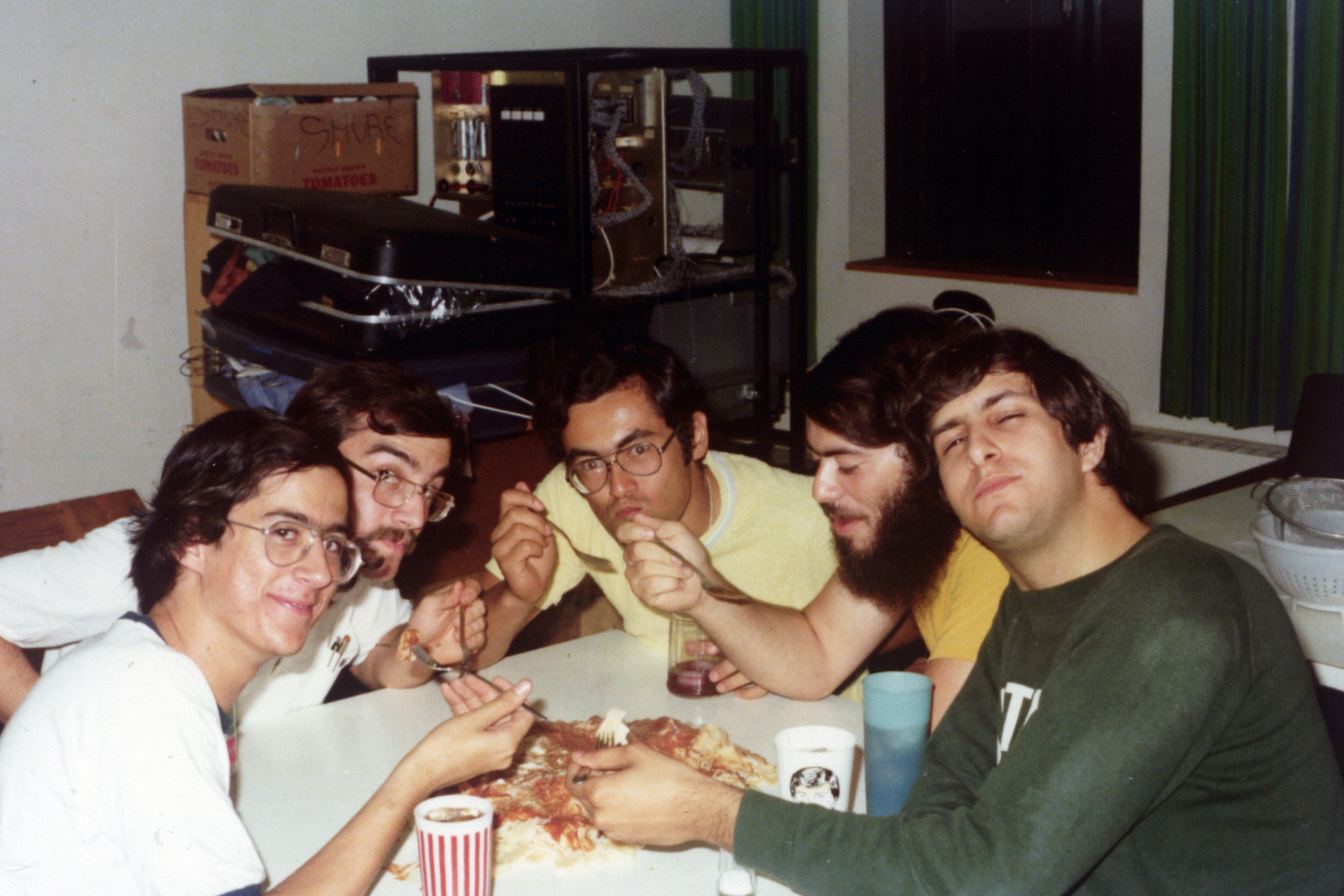 Five men sit around a table eating pizza with forks. There are some cups on the table and a suitcase and some boxes are visible in the background.