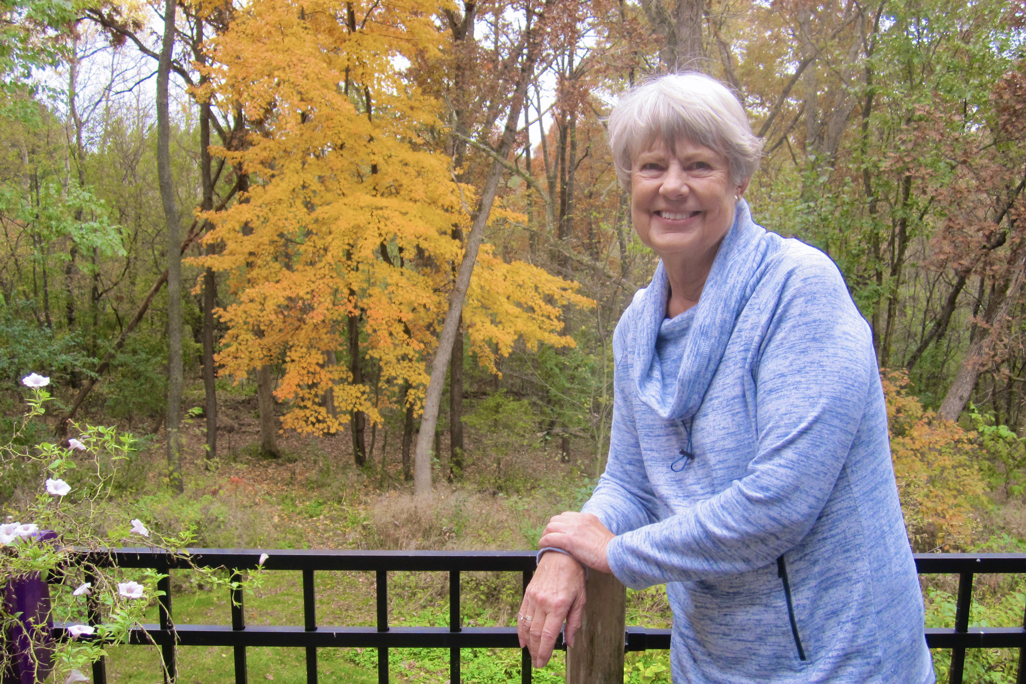 Sara Clope Long is shown facing the camera. She is outside and resting her wrist on a black railing. The area in the background is wooded.