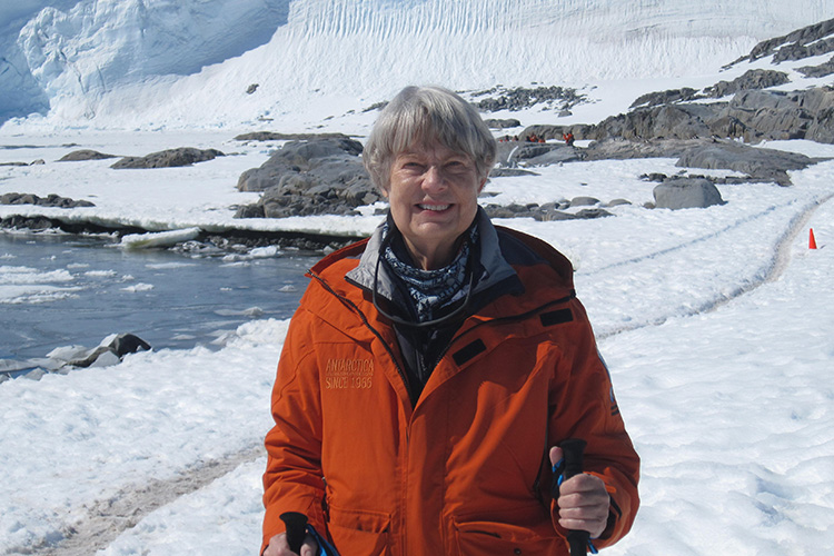 Ara Long is shown from the waist up wearing a red winter coat. A landscape of snow, rocks, and icy water is in the background.