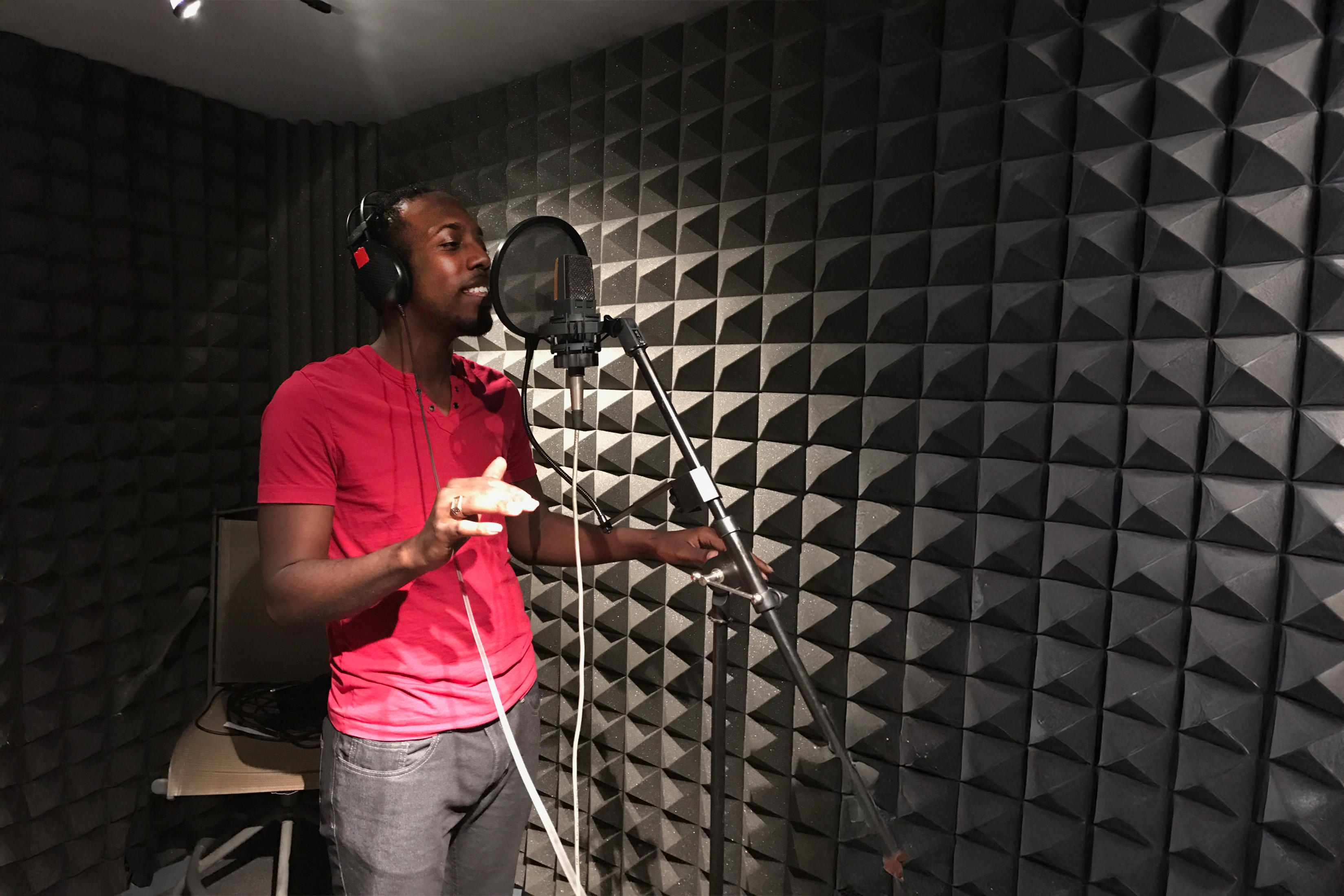 Kelvin Frazier is shown in a recording booth singing into a microphone. The wall shows soundproofing.