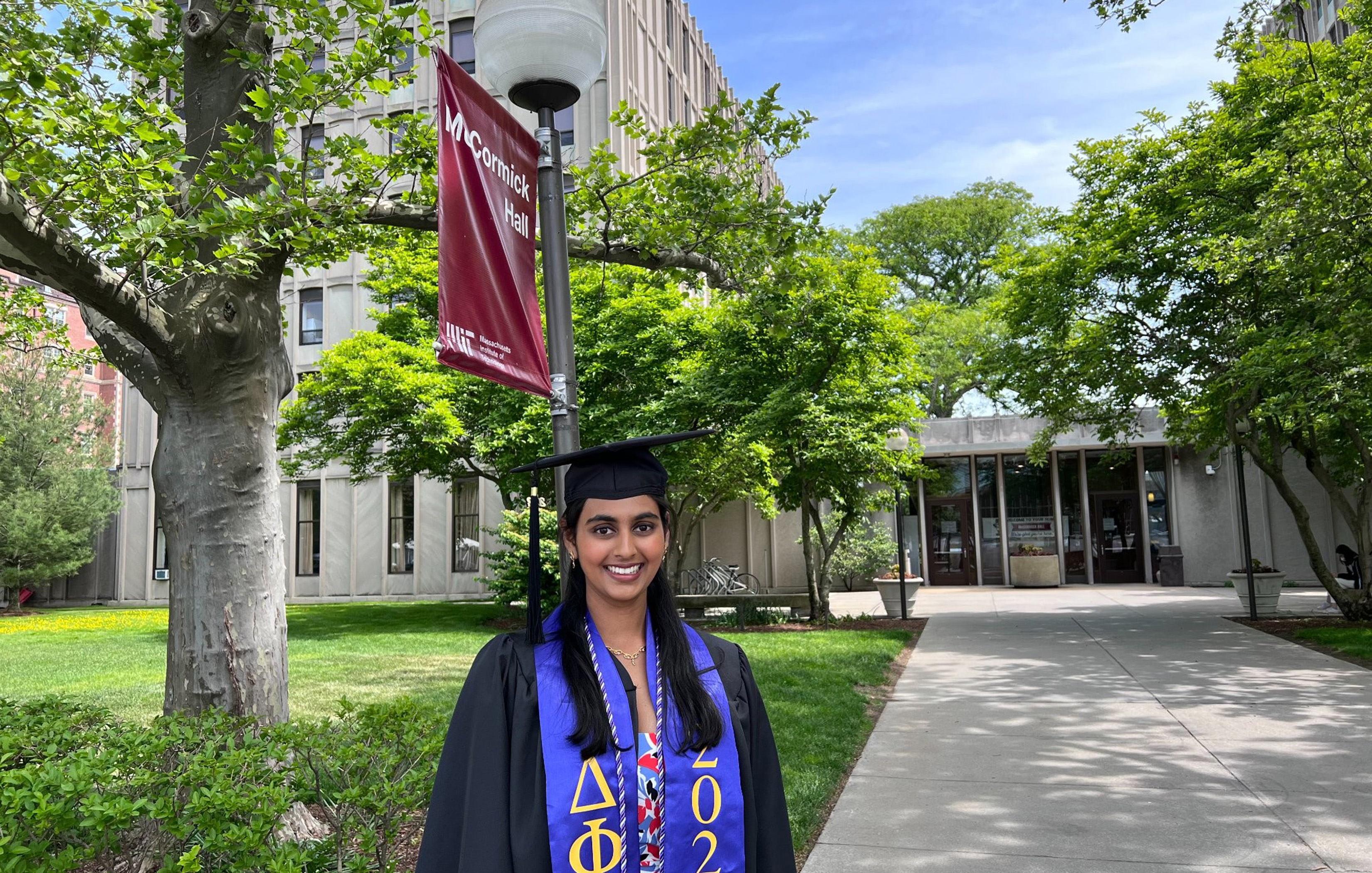 Tara Venkatadri appears in her graduation cap and gown in front of McCormick Hall at MIT. A red banner with the dorm name is attached to a light pole behind her.