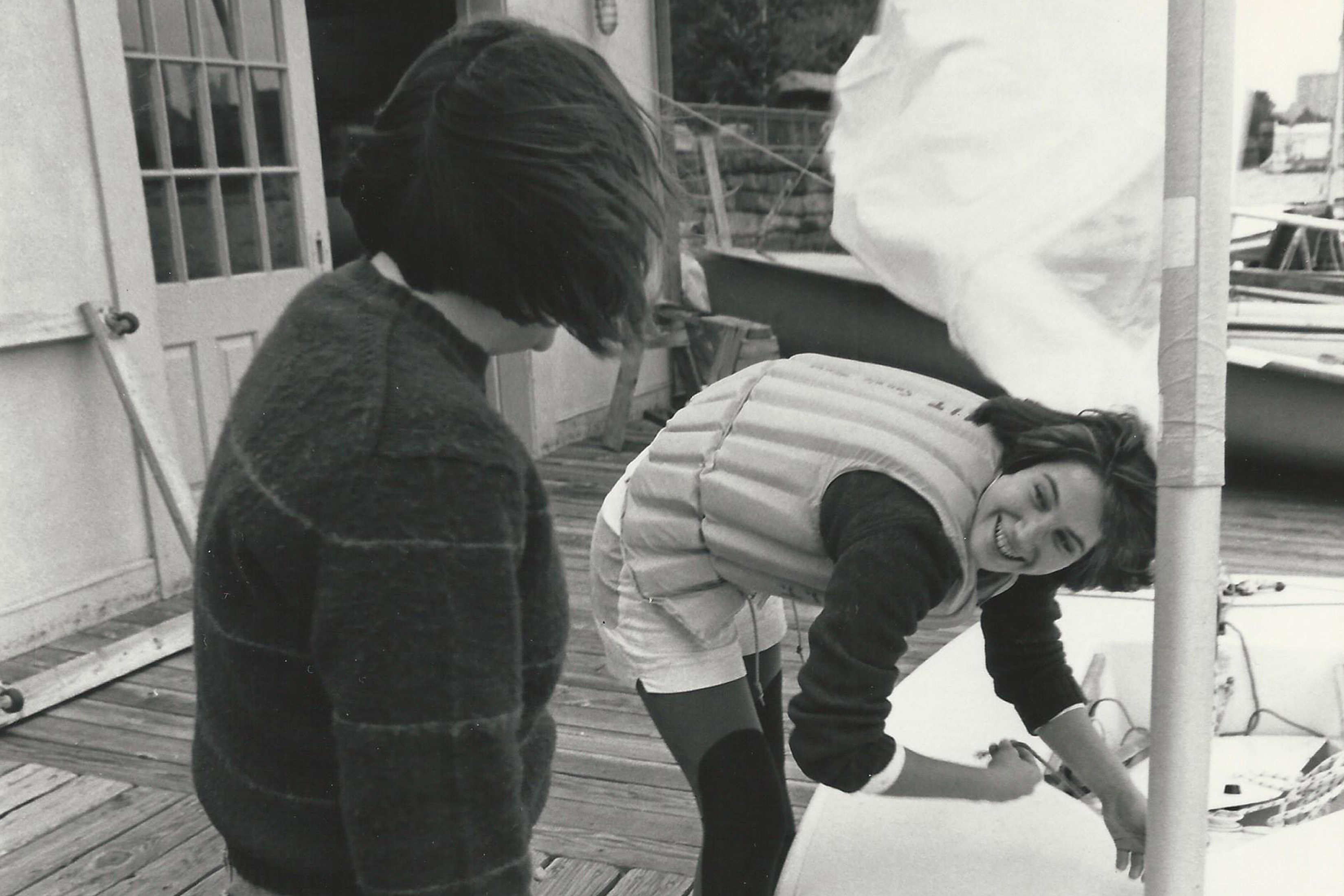 A sailor in a life preserver bends over a boat while looking up at another person whose back is to the camera. A sail flaps in the background.