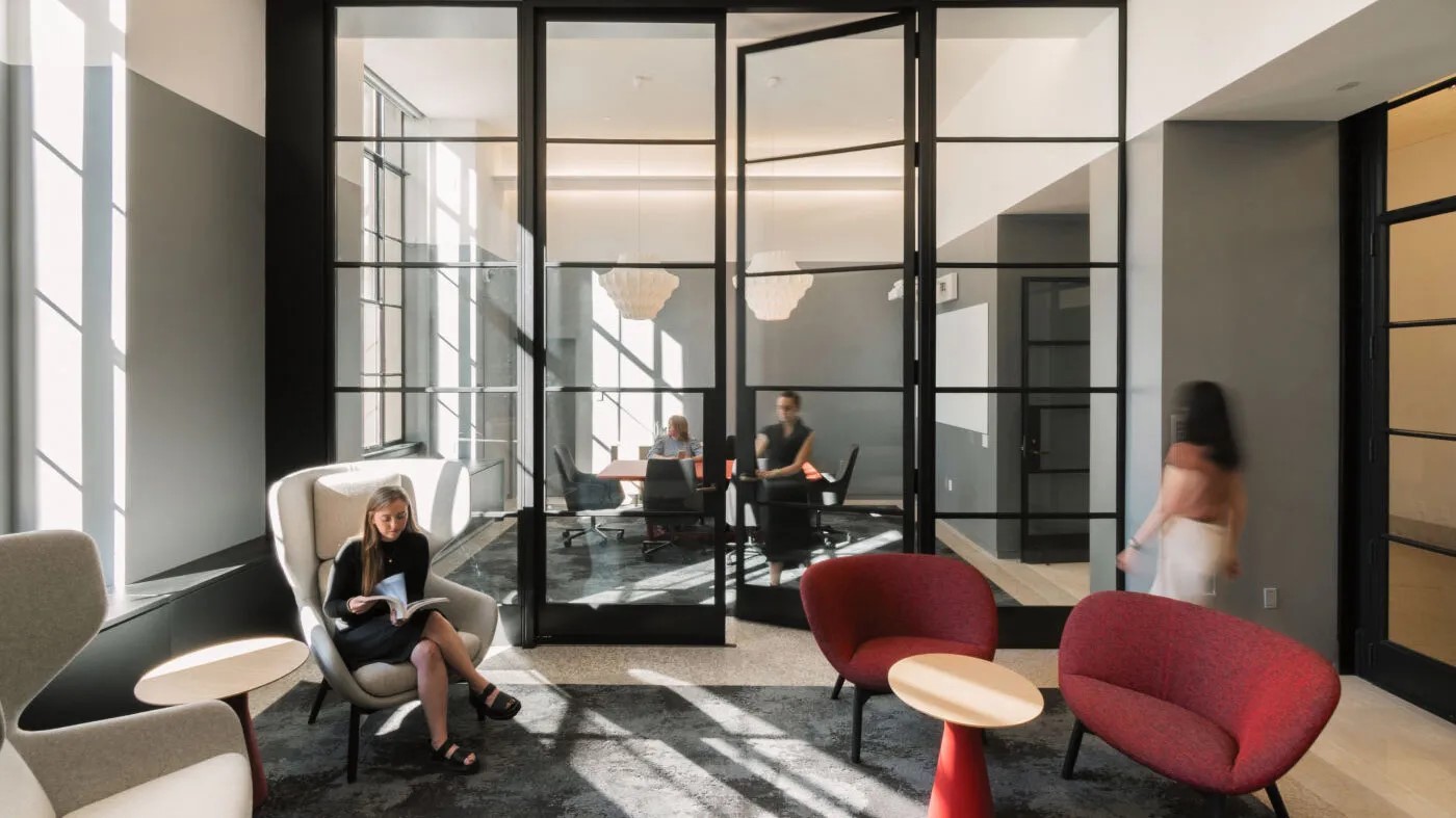 Candid photo of the MIT Alumni Lounge. Person walking to the right past two red chairs. Person sitting and reading to the left on two white chairs. Conference room depicted behind glass french doors