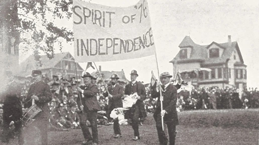 Black and white photo of alumni parading and carrying a sign that reads "Spirit of '76 Independence"