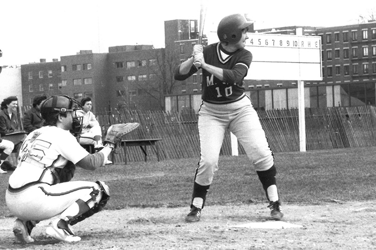 Black and white image shows a softball batter poised to swing, with catcher at left ready to catch. A few women sit on a bench looking on. A blank scoreboard takes up the right corner of the photo, and some buildings are in the background.