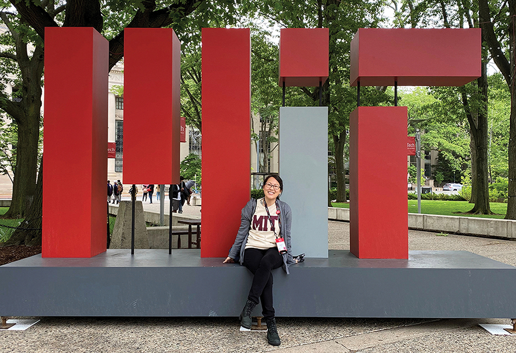 A photo of MIT alum Jeanne Yu sitting on a sign with large "MIT" letters outside with trees and leaves in the background