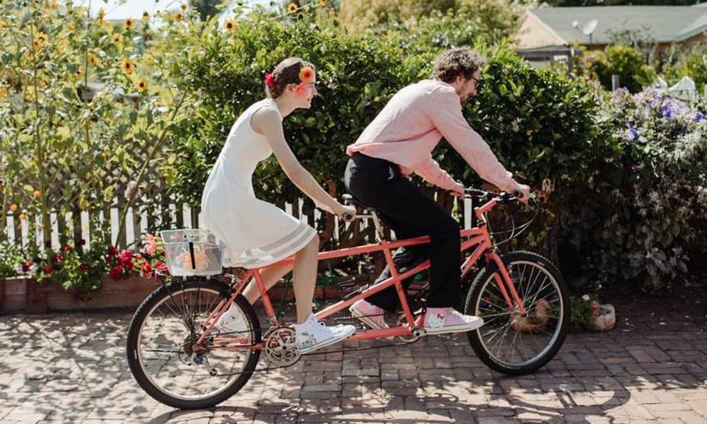Eli Davis and Jenny Ramseyer riding a tandem bicycle on a brick walkway with sunflowers behind them