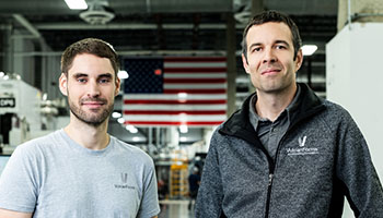 Martin Feldmann in a gray T-shirt and John Hart in a black sweater vest face the camera in an industrial setting. An American flag appears in the background between them.