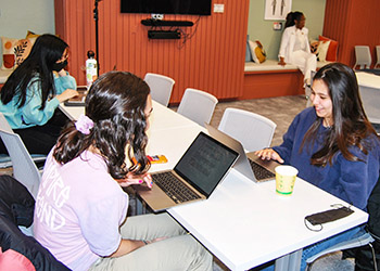 3 women work on laptops sitting at a table. Another table with chairs and a TV screen are in the backgroun