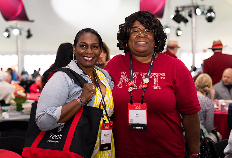 Two women pose in a party tent with MIT gear, including an MIT T-shirt and a reunions tote bag.