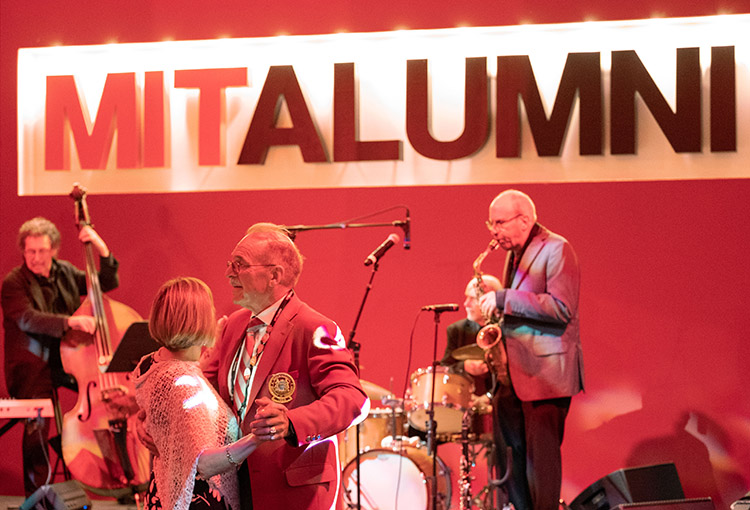 Band members and dancers are shown with a big MIT Alumni banner in the background.