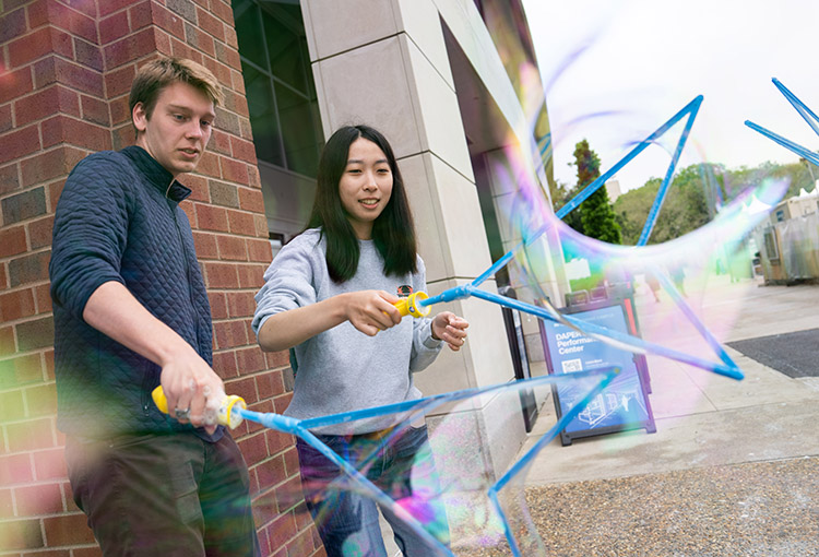 A man and woman form giant bubbles with plastic wands outside.