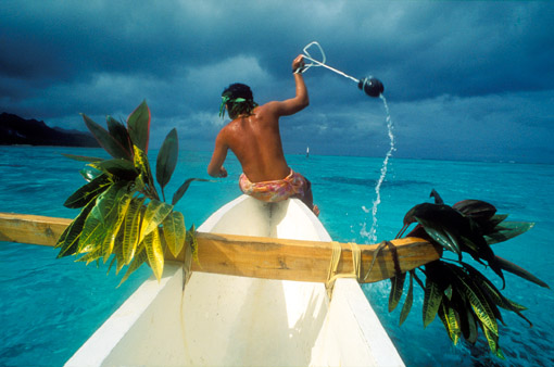 Pêches aux cailloux, fishing with rocks, in Moorea, an island in French Polynesia. Men in boats in a circle smash rocks into the water chasing the fish toward nets near the shore (© Owen Franken).