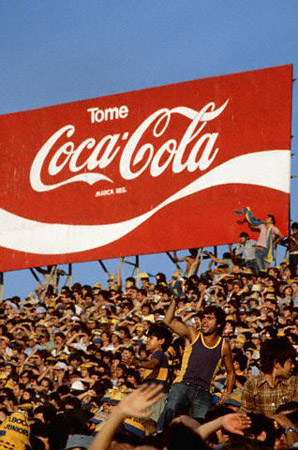 Soccer fans wearing gray and yellow scream and wave their arms for their team under a Coca-Cola billboard in a Buenos Aires stadium, March 1981 (© Owen Franken/CORBIS).