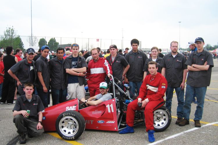 A red race car in a parking lot surrounded by a group of people