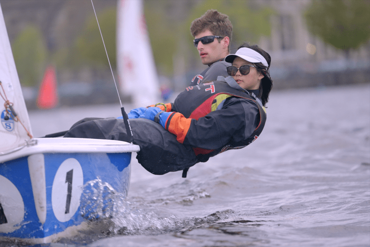 Two MIT students lean out of a small sailboat with their bodies half hanging over the Charles River