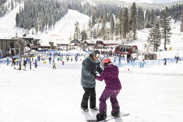 A man helps a woman on a snowboard with a ski resort in the background