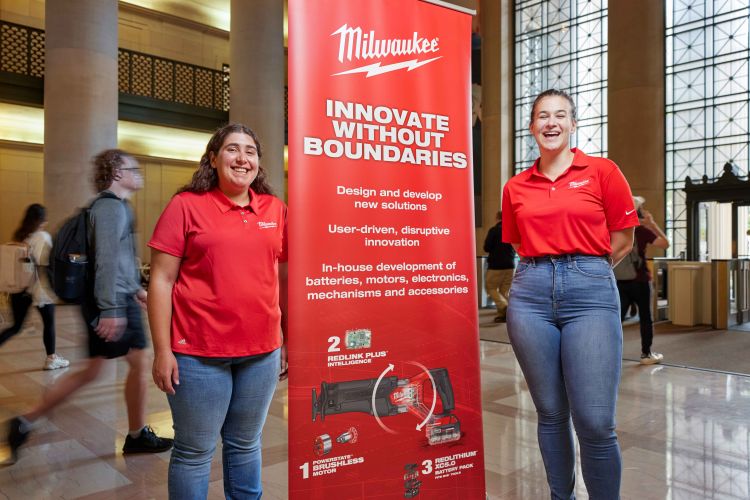 Photo of Beth Cholst (left) and Rosalie Phillips standing in the lobby of Building 7 at MIT, with a red Milwaukee Tool banner in between them and people passing behind them