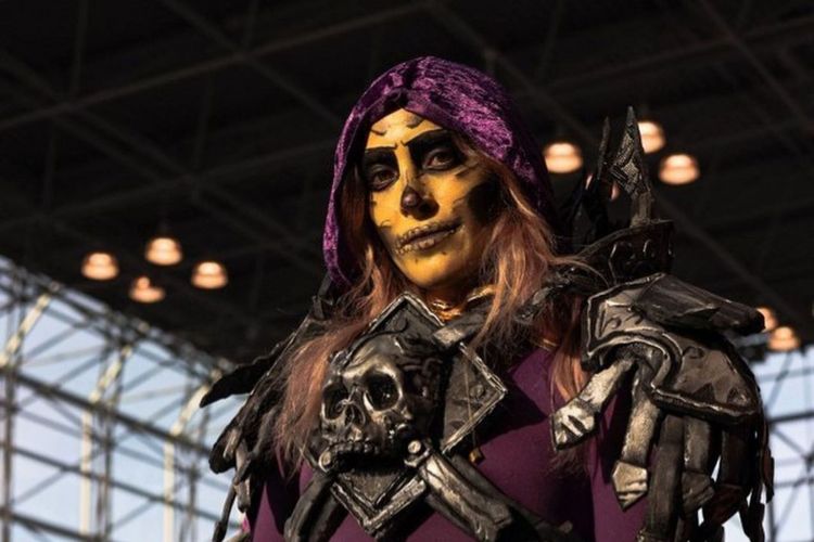 Emma Kay in costume, from chest up. She wears a purple hood, yellowish makeup, and a skull breastplate.