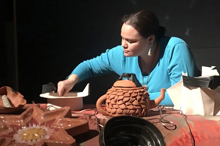 Erin Genia is shown from chest up leaning over an array of clay artwork, including a teapot and a large star.