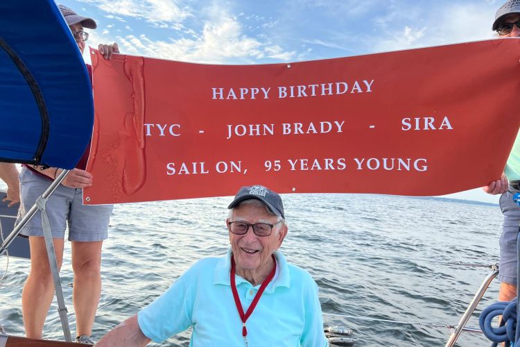 John Brady wears a blue shirt and a baseball cap. A huge red banner held up behind him reads, "Happy birthday - TYC - John Brady - SIRA/ Sail on, 96 Years Young." Water is in the background.