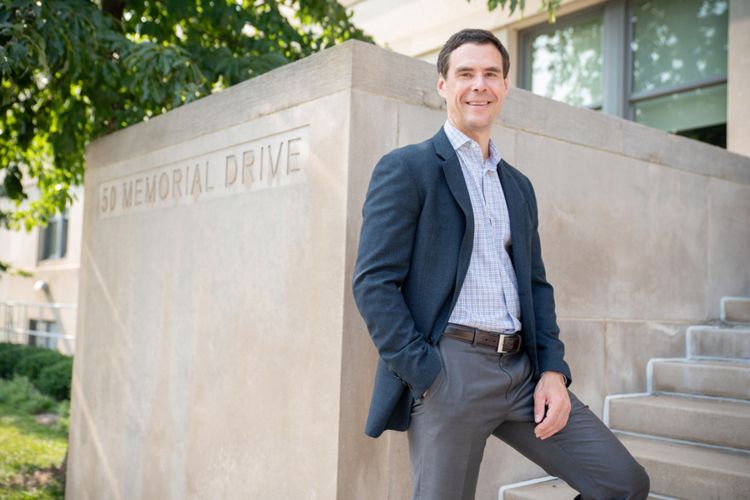 Nathaniel Hendron stands at the bottom of stone steps next to a plinth that reads "50 Memorial Drive." He is wearing a blue blazer, gray slacks, and a button-down shirt.