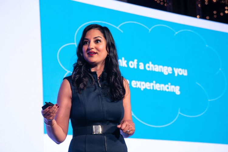 MIT alum Parul Somani holds a clicker standing in front of a big screen where the words "of a change you" and "experiencing" appear in white on a blue background behind her. 