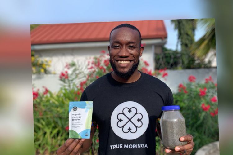 Kwami Williams standing outside with a white building behind him with a red roof, holding True Moringa products and wearing a True Meringa tshirt 