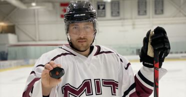 Photo of Bill Near standing in an ice rink wearing an MIT jersey and holding a black plastic device in his right hand and a hockey glove and stick in his left