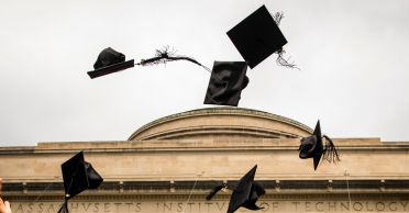 Graduation hats fly through the air against a background of gray sky and the top of the MIT dome