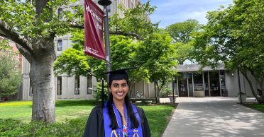 Tara Venkatadri appears in her graduation cap and gown in front of McCormick Hall at MIT. A red banner with the dorm name is attached to a light pole behind her.