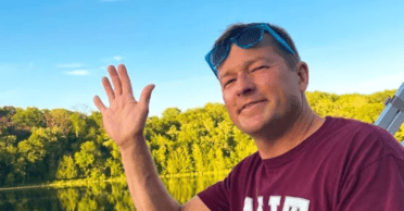 Headshot of Andy Obst ’90 waving while on a boat, sunglasses perched on his head. He's wearing an MIT t-shirt