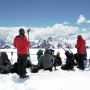 A photo of a crew of people sitting and standing in snow with filming equipment and with mountains in the background