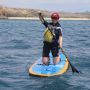 Woman wearing a wetsuit kneeling on a paddleboard in the ocean with a beach and rocky coastline behind her