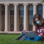 A student in a mask and MIT sweatshirt works on her laptop on the grass in Killian Court